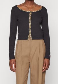 Svart långärmad kortärmad knappar upp topp med leopardmönstrad kant, bärs med högmidjade beigefärgade veckade byxor och tunna guldhalsband.