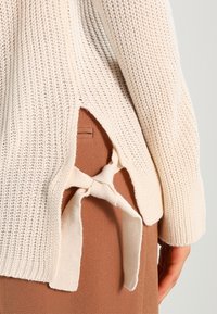 Ljus krämfärgad stickad tröja med avslappnad passform, öppna slynger i sidorna och en knytbar detalj bak med mjuk textur.