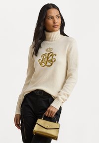 Femeie îmbrăcată cu un pulover crem cu guler înalt, având un monogram în formă de coroana aurie, și pantaloni negri, ținând în mâna dreaptă o mică geantă strălucitoare de aur.
