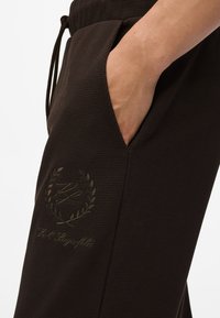 Barna textúrájú nadrág oldalsó zsebbel, sötétebb árnyalatú embosszált logóval, zsinóros derékrésszel és sima anyagú felülettel.