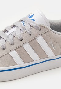 Ljusgrå mockasinsneaker med vita läderränder, blå logodetaljer, runda snören och en texturerad gummisula. Har "CAMPU"-märkning.