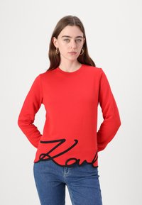 Červená mikina s dlhým rukávom vyrobená z bavlnenej zmesi, s veľkým čiernym kurzívnym logom "Karl" na leme a okrúhlym výstrihom.