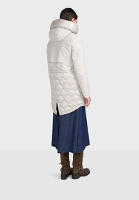 Biely prešívaný kabát s vysokým golierom, kombinujúci lesklé a matné textúry. Výrazný rozparok vzadu a hladký povrch, skombinovaný s džínsovou sukňou a hnedými čižmami.