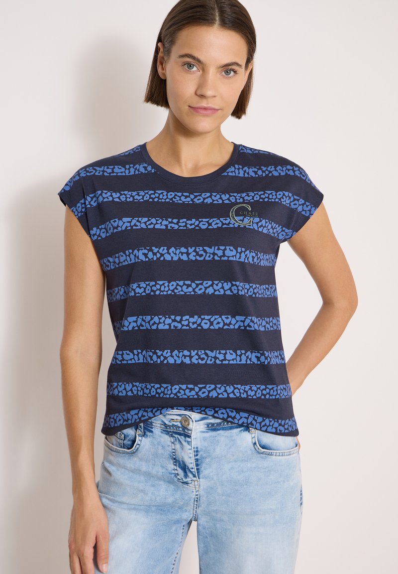 Femme portant une chemise à manches courtes à motifs bleu marine et bleu clair avec un jean bleu clair, debout devant un fond clair uni.