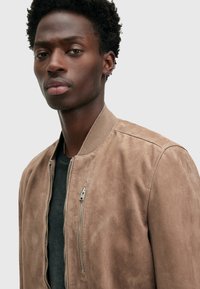 Giacca bomber in suede marrone con zip anteriore, colletto a coste e tasche laterali. Tessuto liscio e dettagli di design minimalisti.