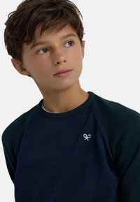 Sudadera azul marino con mangas raglán verde oscuro, cuello acanalado y pequeño logo de tijeras blancas en el pecho. Textura suave.
