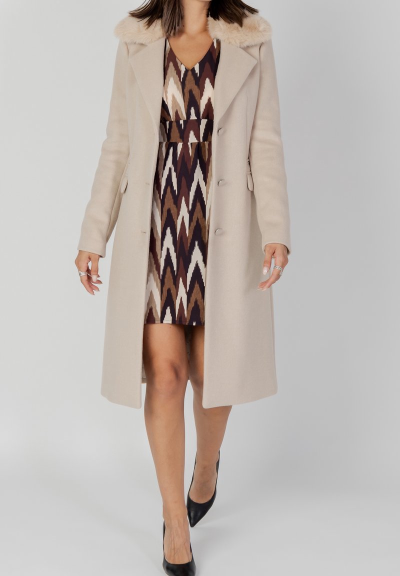 Abrigo de lana beige con cuello de pelo sintético, diseño de botonadura sencilla, bolsillos laterales, usado sobre un vestido estampado en tonos terrosos.