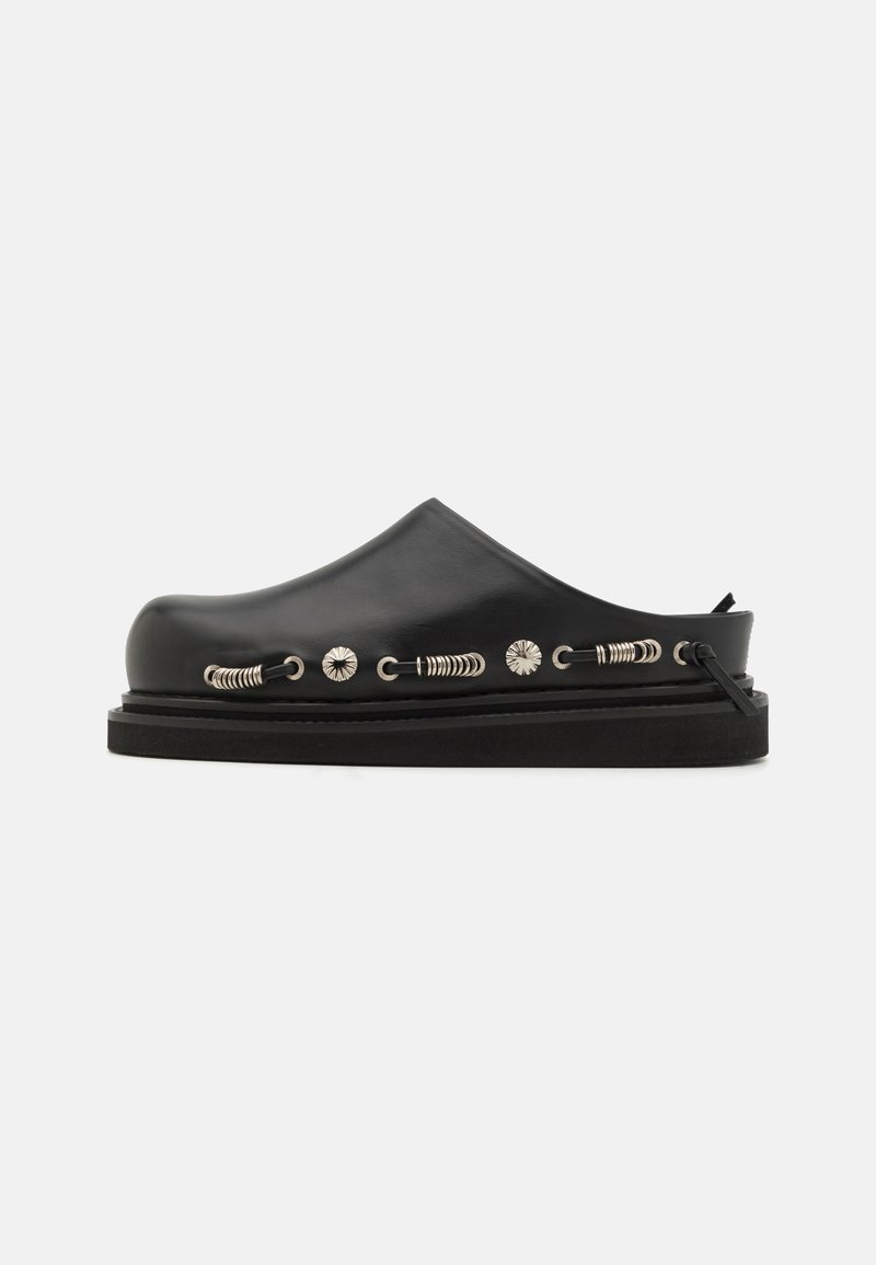 Black leather slide shoe with a rounded toe, textured surface, decorative silver hardware, and a thick rubber sole.