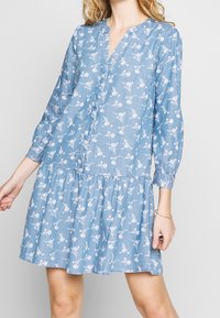 Jasnoniebieska sukienka w kwiatowy wzór, z guzikami z przodu, długimi rękawami i warstwowym dołem. Wykonana z miękkiego materiału w luźnym kroju.