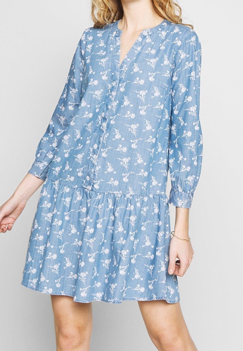 Jasnoniebieska sukienka w kwiatowy wzór, z guzikami z przodu, długimi rękawami i warstwowym dołem. Wykonana z miękkiego materiału w luźnym kroju.