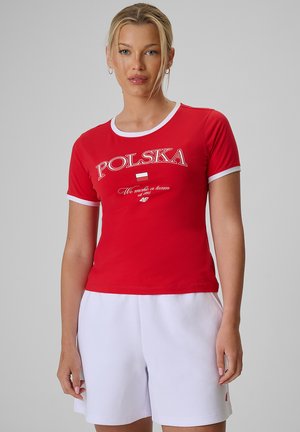 Kobieta w czerwonej koszulce z "POLSKA" i białych szortach na jednolitym tle, stojąca w swobodnej pozie.
