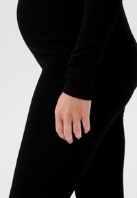 Leggings de maternité noirs à côtes avec un design ajusté, présentant une texture lisse et un gros plan d'une main reposant sur le côté.