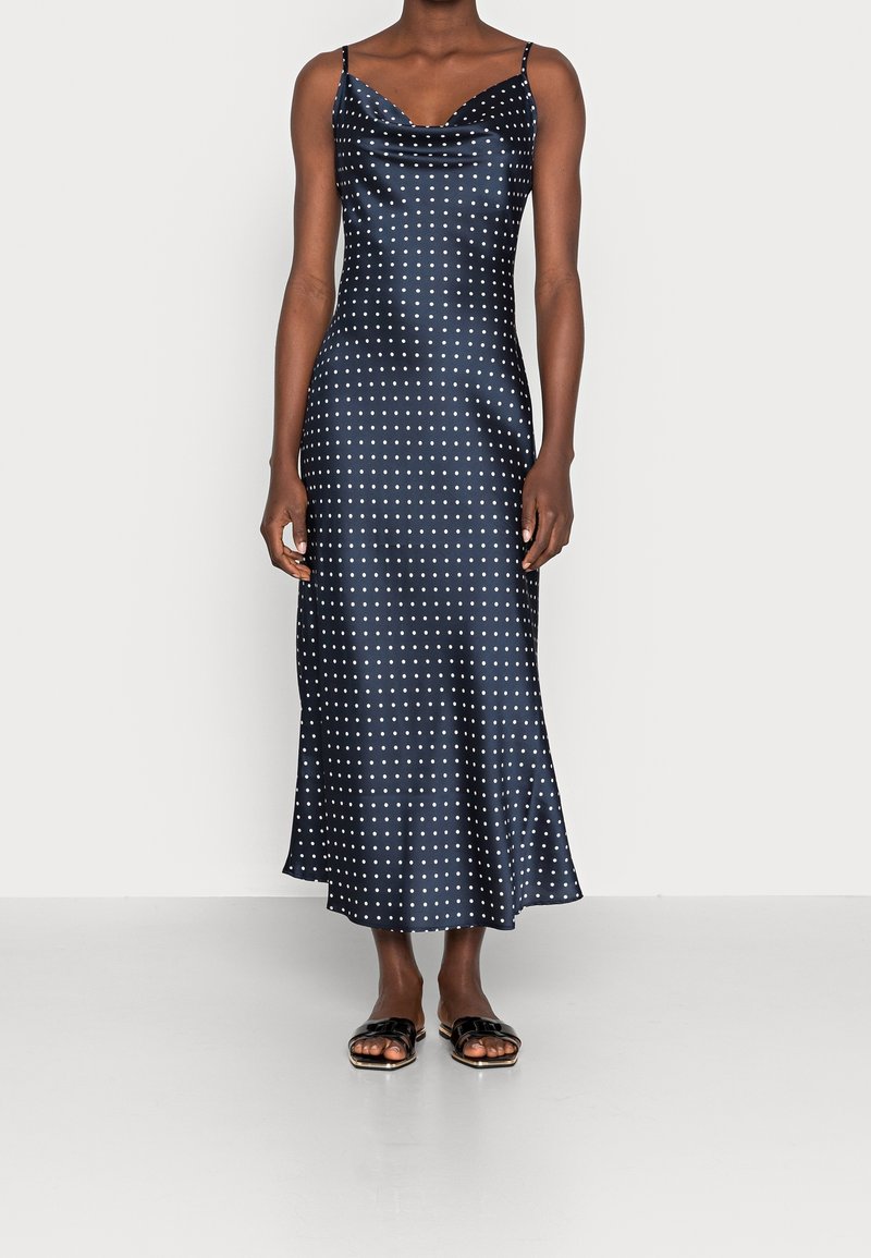 Robe en satin bleu marine à pois, avec un décolleté plongeant, des fines bretelles et un ourlet fluide, associée à des sandales à glisser noires.