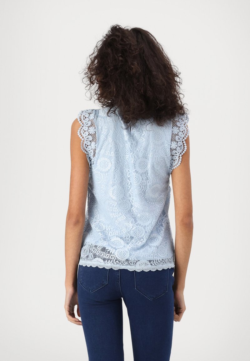 Blouse en dentelle bleu clair avec manches et ourlet festonnés, présentant des motifs floraux complexes et une texture lisse. Portée avec un jean foncé.
