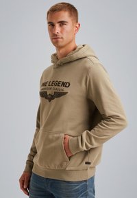 Beige hoodie van zacht stof, voorzien van een voorzak in kangoeroestijl, een capuchon met trekkoorden en een bedrukte logo met vleugels op de borst.