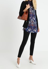 Svart långärmad blazer, mönstrad blå tunika med blommönster, svarta leggings och vita spetsiga klackar, kompletterat med en brun handväska.