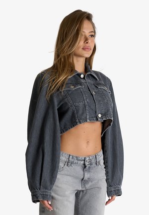 Giovane donna che indossa una giacca di jeans nera corta e jeans grigio chiaro a vita alta, in piedi davanti a uno sfondo bianco semplice.