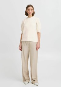 Top en tricot texturé écru avec des manches courtes bouffantes, associé à un pantalon large beige clair et des chaussures blanches à bouts pointus.