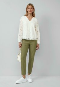Blouse en dentelle blanche avec un décolleté en V, des manches longues et des détails en œillets. Pantalons slim-fit vert olive et baskets blanches. Petit sac à main blanc avec une chaîne.