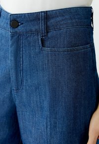 Dunkelblaue Jeanshose mit flachem Bund, Gürtelschlaufen, einem Knopfverschluss und einer Seitentasche. Strukturiertes Material mit einem dezenten Glanz.