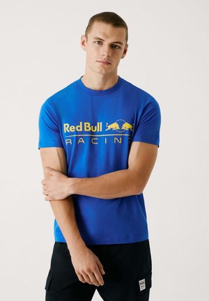 Camiseta de manga corta azul de algodón, con un logo amarillo de "Red Bull Racing" y una textura suave. Combinada con pantalones negros.