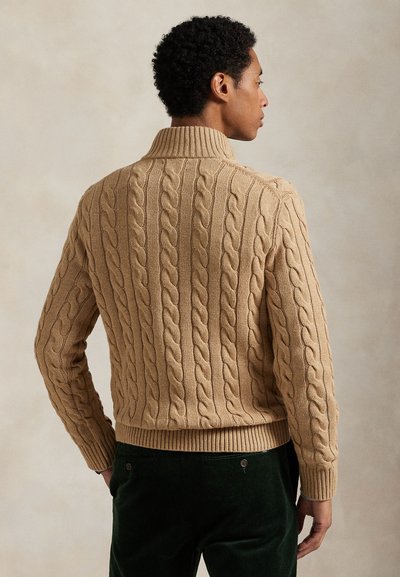 Persoon van achteren gezien die een beige kabelgebreide trui met hoge kraag en een donkere broek draagt tegen een effen achtergrond.