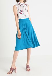 Blouse florale blanche à col haut, associée à une jupe plissée teal. La modèle porte des sandales à talons carrés roses. Tissu lisse, contraste de couleurs vives.