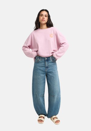 Femme aux longs cheveux foncés portant un sweat-shirt rose ample, un jean bleu large et des sandales blanches, debout les mains sur les hanches.
