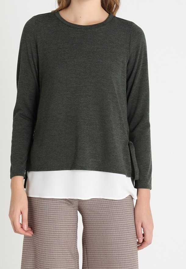 Haut à manches longues vert foncé en tissu texturé, avec un col arrondi et des fentes sur les côtés, superposé sur une chemise blanche.