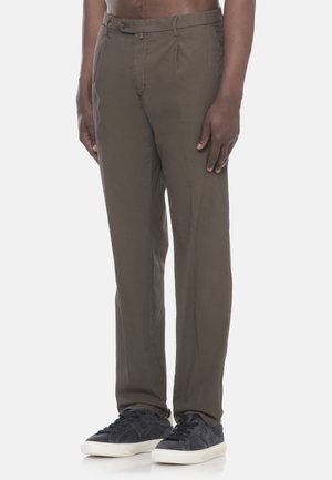 Uomo che indossa pantaloni marroni a gamba dritta e sneakers nere con suola bianca, in piedi davanti a uno sfondo chiaro uniforme.
