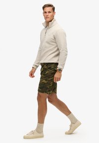 Sudadera de cremallera color beige claro, hecha de material suave, combinada con pantalones cortos cargo de camuflaje verde. Zapatillas color beige y calcetines beige completan el conjunto.