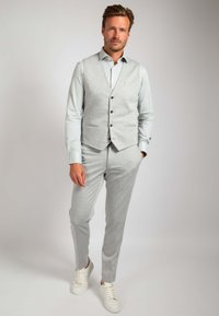 Hellgrauer dreiteiliger Anzug mit einer strukturierten Weste, passender Hose und einem hellblauen Hemd. Akzentuiert mit braunen Knöpfen und weißen Sneakers.