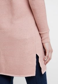 Maglione lavorato a maglia rosa chiaro con texture a coste, aperture laterali e polsini aderenti, indossato con jeans scuri.