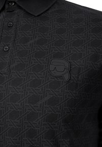 Svart pikétröja med ett diskret geometriskt mönster. Inkluderar en texturerad svart logotypmärke med en stiliserad ansiktsdesign på vänster bröst.