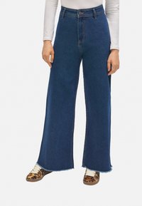 Jeans de tiro alto y pierna ancha en denim azul oscuro con dobladillo sin rematar. Combinados con zapatillas de estampado animal, que tienen cordones en beige y suelas texturizadas.