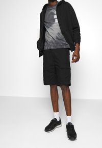 Haut à zip noir, t-shirt gris tie-dye, shorts cargo noirs et baskets noires avec des chaussettes blanches. Design athlétique simple, accents minimaux.