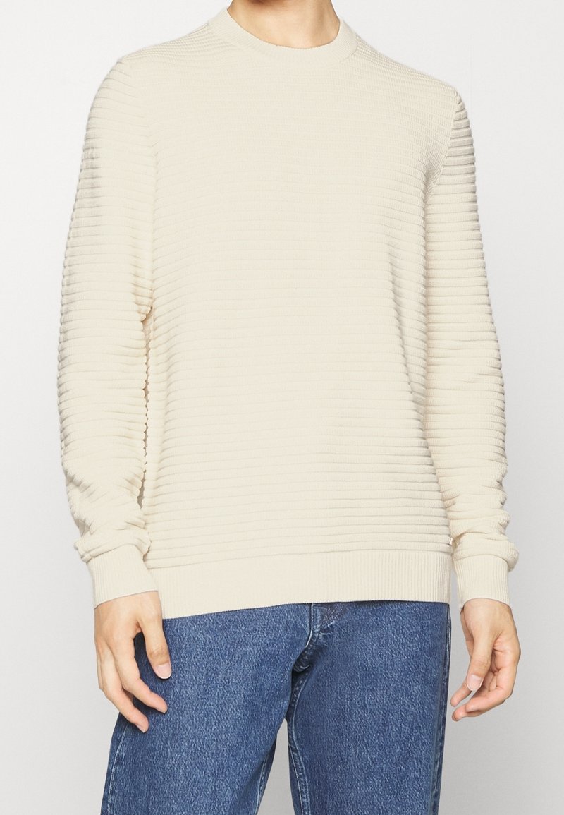 Personne portant un pull crème à manches longues côtelé et un jean bleu, les mains détendues le long du corps, sur un fond clair uni.