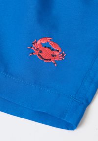 Rød broderet krabbe på kanten af lyst blåt stof, der viser detaljerede sting og rene sømlinjer.