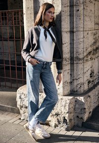 Modelka nosící tmavě šedou bomber bundu, bílé tričko, světle modré džíny s rovnými nohavicemi a krémové tenisky s béžovými doplňky, opírající se o kamennou zeď.