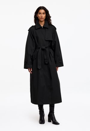 Femme portant un long trench-coat noir ceinturé et des bottes noires à talons, debout devant un fond blanc uni.