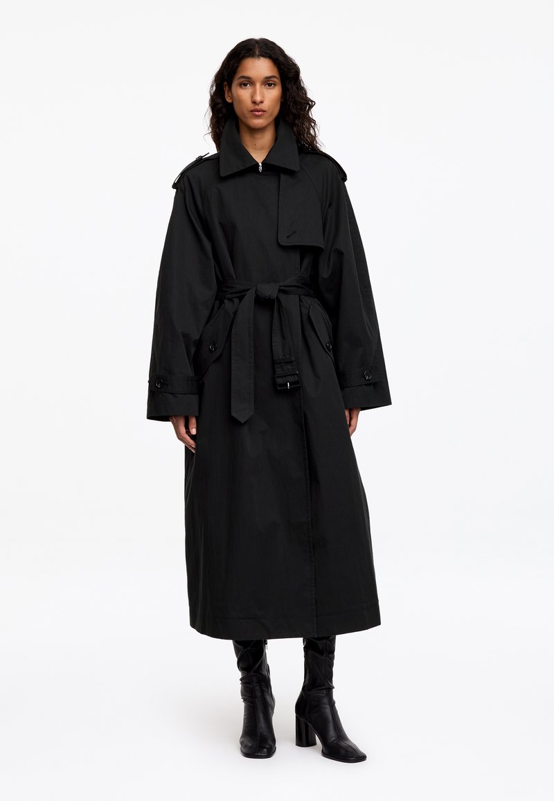 Femme portant un long trench-coat noir ceinturé et des bottes noires à talons, debout devant un fond blanc uni.