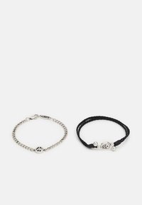 Duas pulseiras: uma pulseira de corrente de prata com um símbolo da paz e uma pulseira de corda preta trançada com uma caveira de prata e contas.
