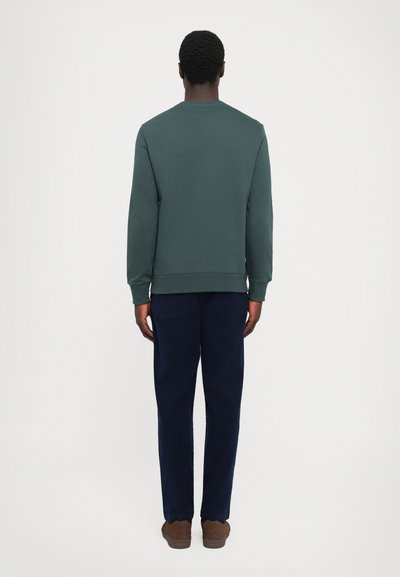 Hombre de pie de espaldas, vistiendo un sudadera verde oscuro, pantalones azul marino y zapatos marrones contra un fondo claro y liso.