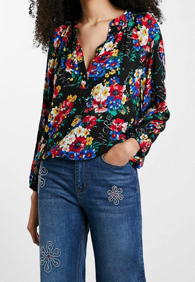 Femme portant un chemisier noir à manches longues avec un imprimé floral coloré et un jean bleu décoré de motifs floraux en strass blancs et rouges.