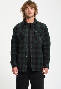 Green and black checkered flannel shirt with long sleeves, snap closure, two front pockets, and a soft, textured fabric. Black t-shirt underneath.