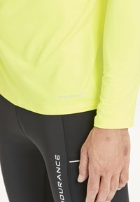 Langærmet, lys gul atletisk skjorte med fugttransporterende stof, parret med sorte leggings med hvid tekst og detaljer.