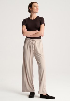 Kobieta stoi z założonymi ramionami, ubrana w dopasowaną czarną koszulkę, beżowe szerokie spodnie z sznurkiem i czarne wsuwane buty.