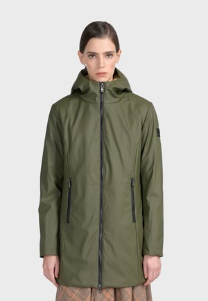 Femme portant un long imperméable à capuche vert olive avec une fermeture éclair à l'avant et des poches zippées sur un fond clair uni.