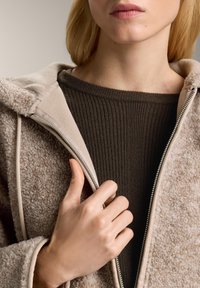 Beżowa polarowa kurtka o teksturowanej powierzchni, z beżowym zamkiem błyskawicznym i brązowym swetrem z prążkami pod spodem. Ręka modela trzyma zamek lekko otwarty.