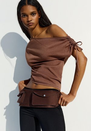 Fiatal nő barna váll nélküli fodros felsőt és hozzá illő barna övtáskát visel, több rekesszel, egyszerű, világos háttér előtt.